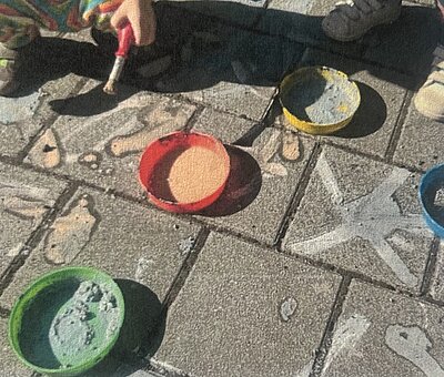 Bunte Kreidefarben in kleinen Schalen stehen auf einem gepflasterten Boden; daneben sind bereits gemalte Kreidemotive und eine Hand mit Pinsel zu sehen.