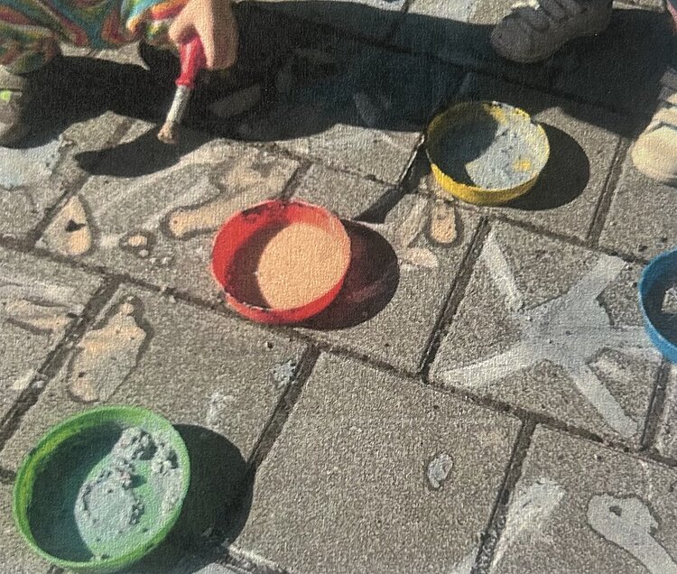 Bunte Kreidefarben in kleinen Schalen stehen auf einem gepflasterten Boden; daneben sind bereits gemalte Kreidemotive und eine Hand mit Pinsel zu sehen.