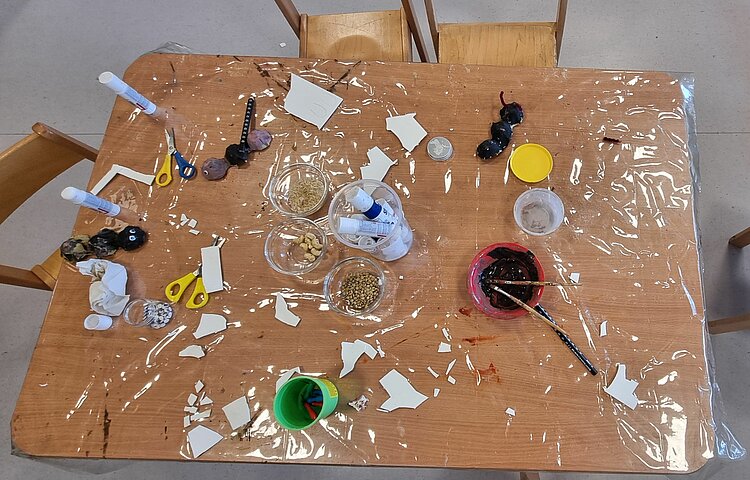 Auf einem mit Plastikfolie abgedecktem Holztisch liegen zahlreiche Bastelmaterialien: Scheren, Papier, Pinsel, Farbe, Klebestifte, und dazwischen gebastelte Ameisen.