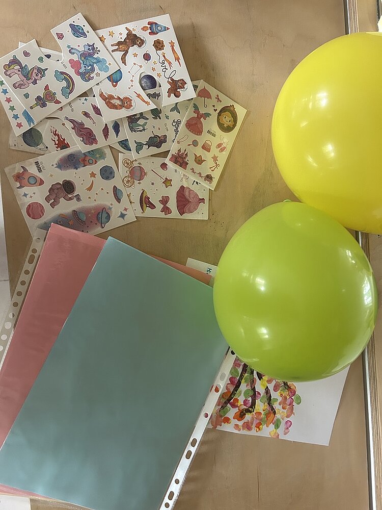 Zwei gelbe Luftballons liegen auf einer Tischplatte. Daneben befinden sich mehrere bunte Stickerbögen mit Motiven wie Meerestieren, Planeten und Fantasiefiguren. Im Vordergrund liegen farbige Papierbögen in Rosa und Hellblau sowie eine Klarsichthülle. Ein weiteres Blatt mit bunten Fingerabdrücken ist teilweise sichtbar.