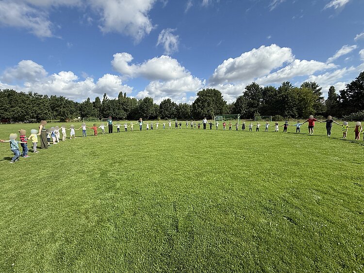 Sportfest der 4- bis 6-jährigen Kinder Eine große Gruppe von Kindern und Erwachsenen steht Hand in Hand in einem Halbkreis auf einer grünen Wiese, unter blauem Himmel mit weißen Wolken.