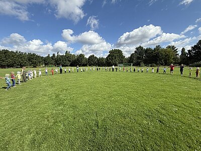 Sportfest der 4- bis 6-jährigen Kinder Eine große Gruppe von Kindern und Erwachsenen steht Hand in Hand in einem Halbkreis auf einer grünen Wiese, unter blauem Himmel mit weißen Wolken.