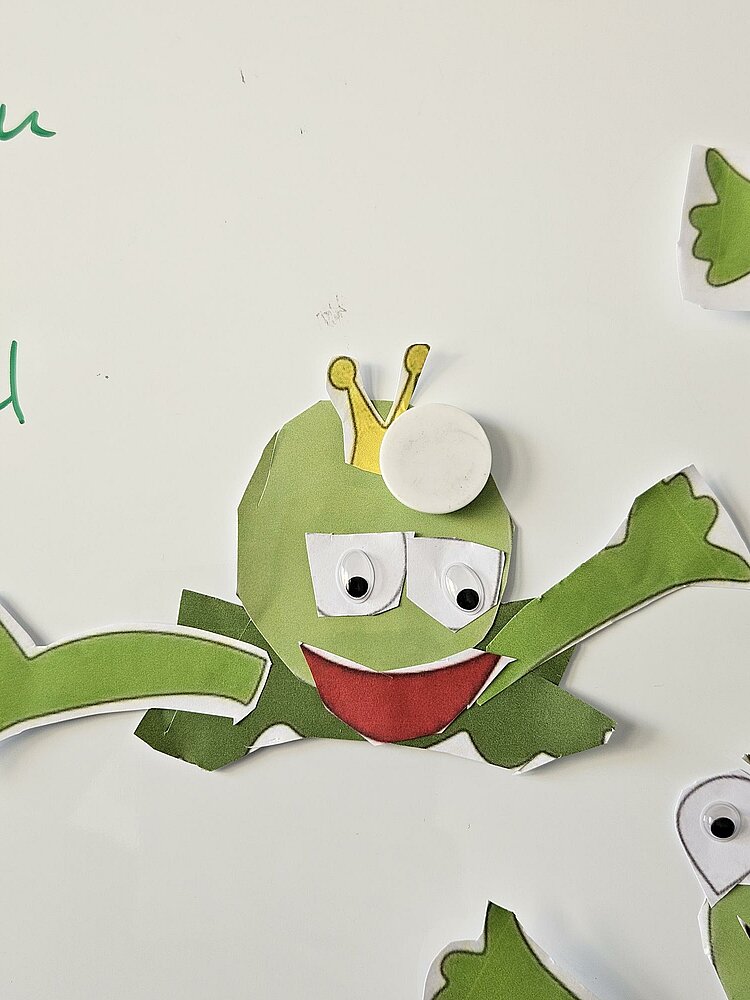 Ein grüner Frosch aus Pappe mit großen Augen, einer Krone auf dem Kopf und einem breiten Lächeln. Der Frosch ist in Einzelteile zerschnitten und auf einem weißen Hintergrund verteilt.