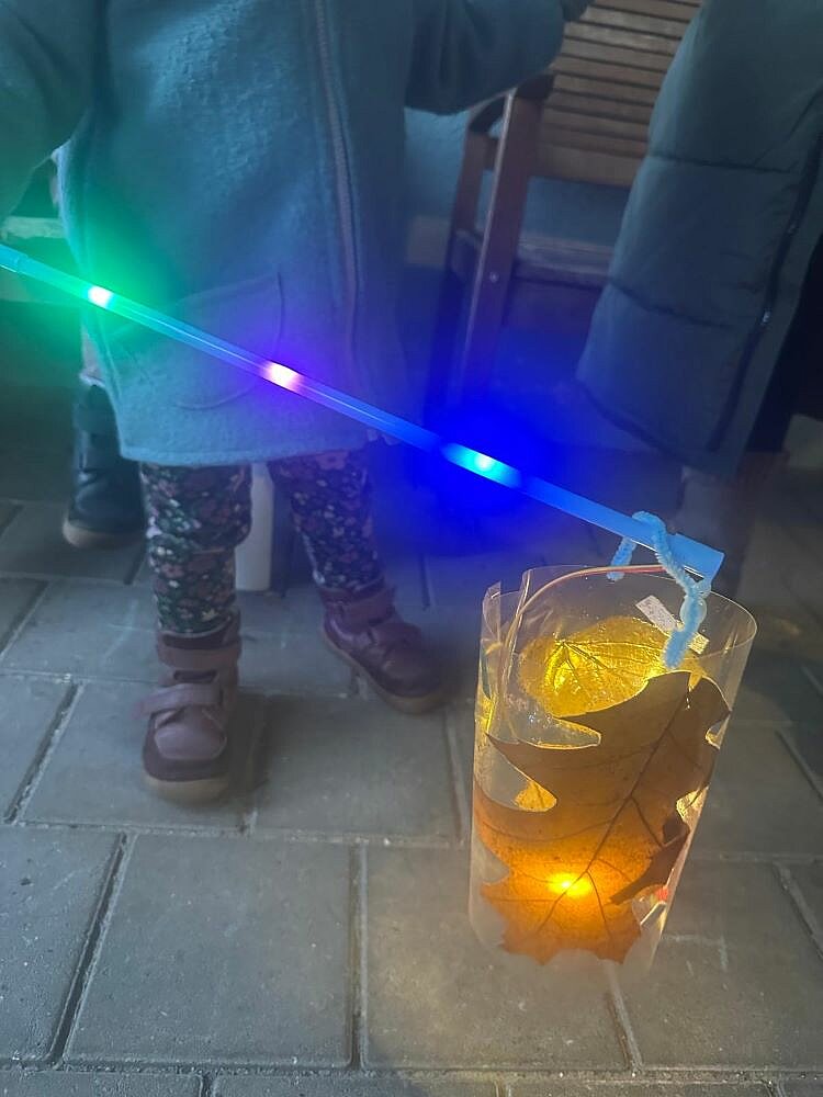 Ein Kind hält einen leuchtenden Stab mit blauen, grünen und violetten Lichtern in der Hand. Daran hängt eine selbstgebastelte Laterne aus durchsichtigem Material, die mit gelben Herbstblättern und einem kleinen LED-Licht dekoriert ist. Das Kind trägt eine Jacke, eine gemusterte Hose und lilafarbene Schuhe.