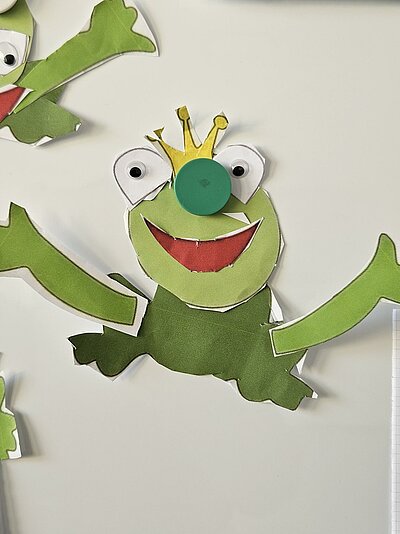 Ein grüner Frosch aus Pappe mit großen Augen, einer Krone auf dem Kopf und einem breiten Lächeln. Der Frosch ist in Einzelteile zerschnitten und auf einem weißen Hintergrund verteilt.
