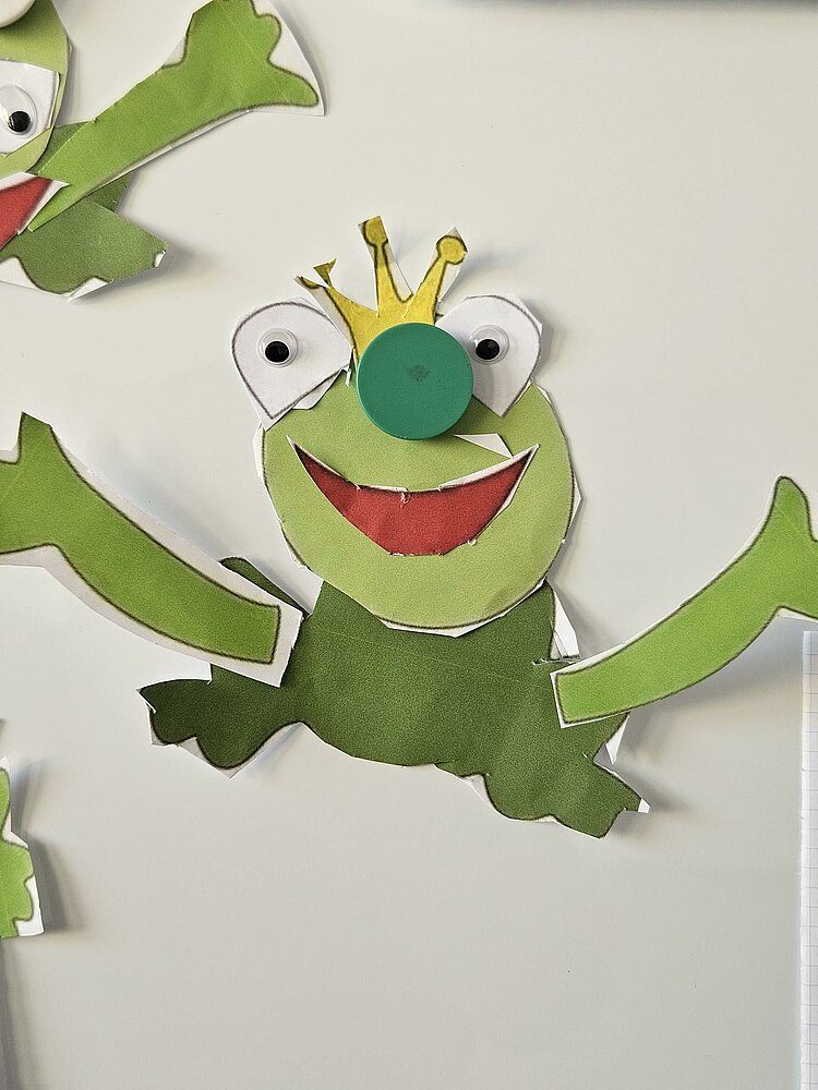 Ein grüner Frosch aus Pappe mit großen Augen, einer Krone auf dem Kopf und einem breiten Lächeln. Der Frosch ist in Einzelteile zerschnitten und auf einem weißen Hintergrund verteilt.