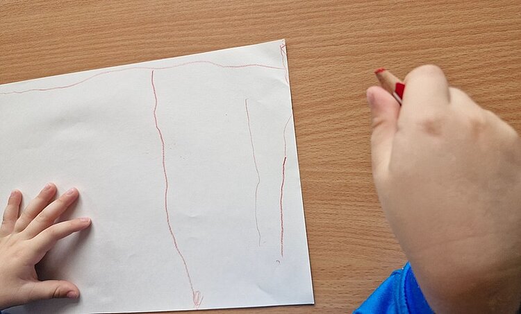 Kinderhand mit blauem Ärmel hält einen roten Buntstift und zeichnet auf einem weißen Blatt Papier, das auf einem hellbraunen Holztisch liegt. Auf dem Papier sind mehrere einfache, senkrechte Linien in Rot und Blau zu sehen. Die andere Hand liegt flach auf dem Papier.
