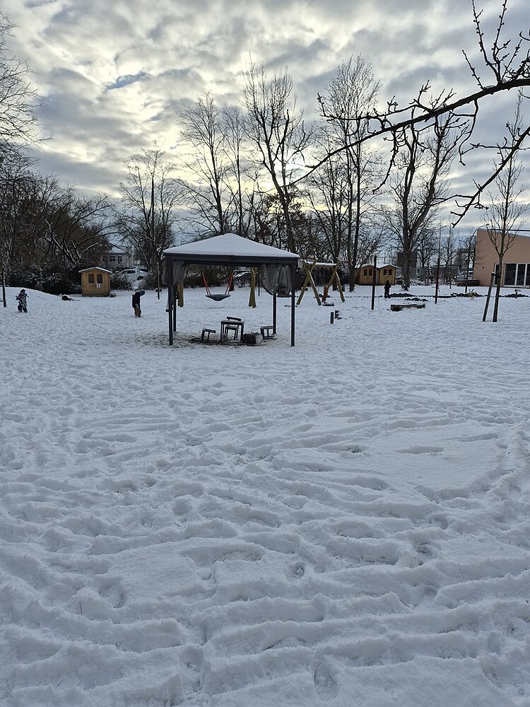 Schneebedeckter Spielplatz mit Pavillon, mehreren Holzhäuschen und kahlen Bäumen im Hintergrund.