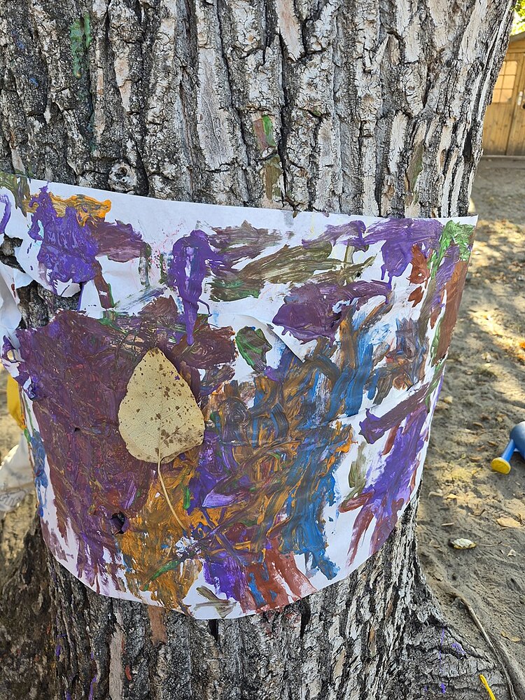 Ein breiter Papierstreifen ist um den Stamm eines Baumes gelegt und mit bunten, abstrakten Pinselstrichen in Lila, Orange, Braun, Grün und Blau bemalt. Ein trockenes Blatt klebt auf dem Papier. Im Hintergrund sind sandiger Boden, ein Zaun und unscharfe Spielgeräte zu erkennen.