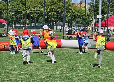 Festspiele mit Ritter Keule beim 1. FC Union Berlin Eine Gruppe von Kindern in bunten Sicherheitswesten und Sonnenhüten läuft auf einem Sportfeld.