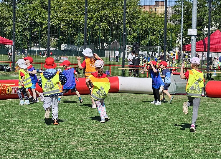 Festspiele mit Ritter Keule beim 1. FC Union Berlin Eine Gruppe von Kindern in bunten Sicherheitswesten und Sonnenhüten läuft auf einem Sportfeld.