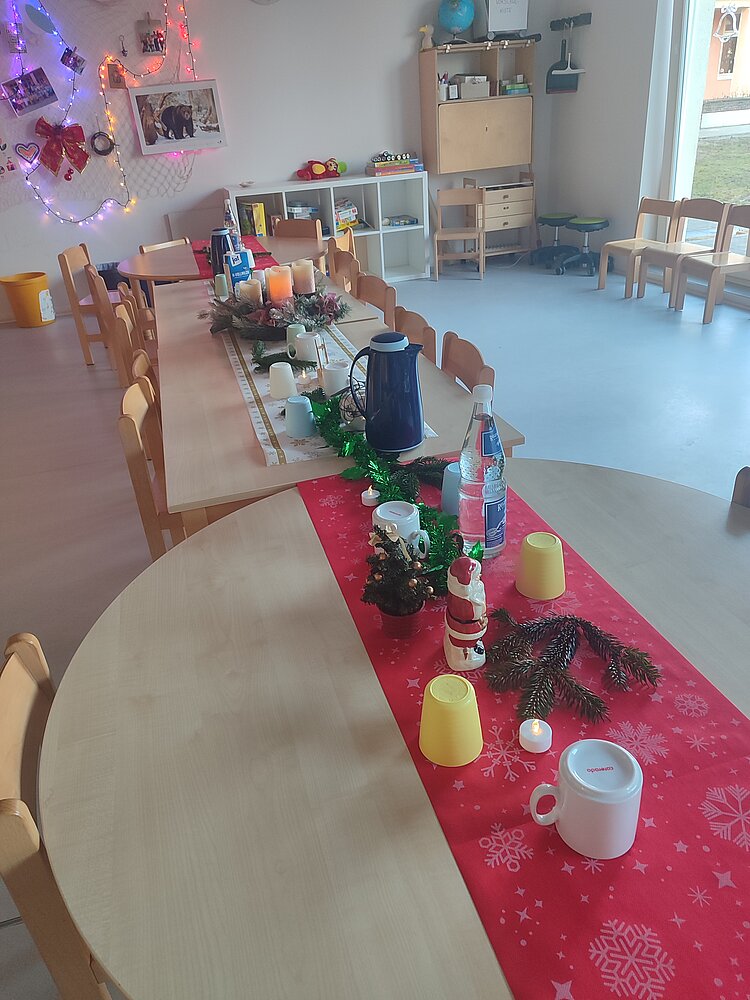 Langer Tisch in einem hellen Gruppenraum, weihnachtlich gedeckt mit rotem Läufer, Tannenzweigen, Kerzen, Bechern und kleinen Dekofiguren.