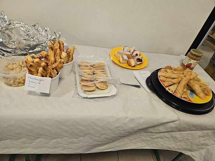 Snack-Buffet mit Käsegebäckstangen, verpackten Keksen, kleinen mit Puderzucker bestäubten Teilchen und einem Teller mit gerollten Crêpes; daneben stehen Löffel und ein Marmeladenglas.