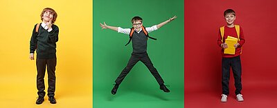 Drei Schulkinder stehen jeweils vor unterschiedlich farbigen Hintergründen. Links steht ein Kind mit lockigen Haaren und Schulranzen vor einer gelben Wand, die Hand am Mund. In der Mitte springt ein Kind mit ausgestreckten Armen und Beinen vor einer grünen Wand, ebenfalls mit Schulranzen. Rechts steht ein Kind mit kurzen Haaren und rotem Pullover vor einer roten Wand und hält Hefte oder Bücher im Arm.