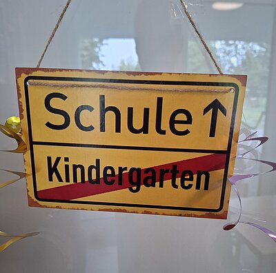 Abschiedsfest der Zaunkönige und Möwen Ein gelbes Schild im Stil eines Ortseingangsschilds hängt an einer Tür. Das Wort ‚Kindergarten‘ ist durchgestrichen, darüber steht in großer Schrift ‚Schule‘ mit einem Pfeil nach oben – ein symbolischer Hinweis auf den Übergang zur Schule.