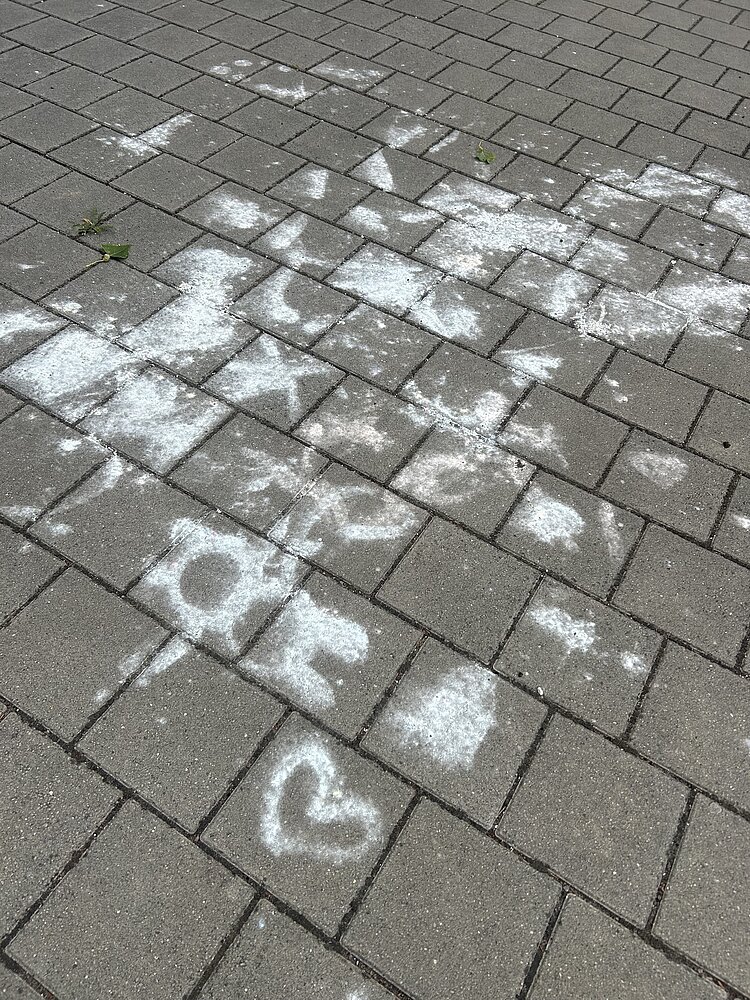 Mit weißer Kreide gestaltete Zeichnungen und Schriftzüge auf grauem Pflaster, darunter Herzen, Symbole und verschiedene Formen.
