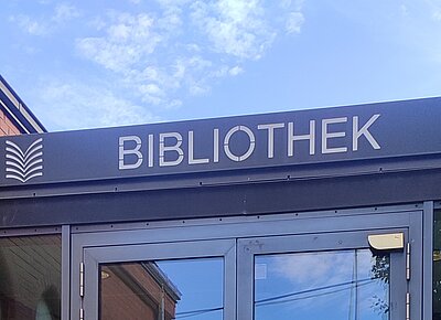Über einem modernen Eingangsbereich befindet sich ein Schild mit dem Wort 'BIBLIOTHEK' in großen, klaren Großbuchstaben. Links daneben steht ein stilisiertes Symbol, das an ein aufgeschlagenes Buch erinnert. Die Fassade besteht aus Glas und Metall, im Hintergrund ist blauer Himmel mit einigen Wolken zu sehen.