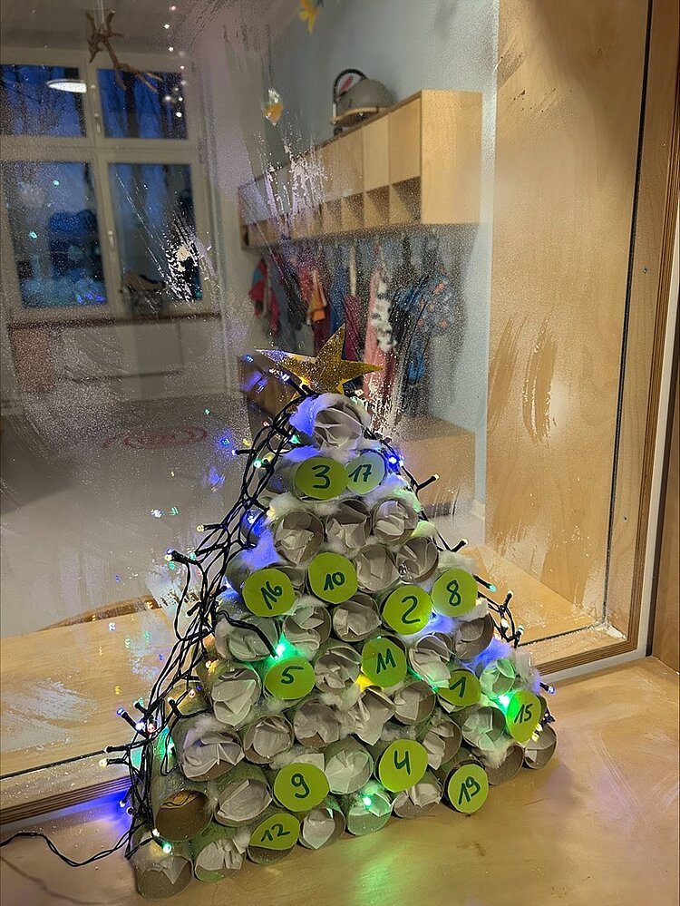 Ein selbst gebastelter Adventskalender in Form eines Dreiecks, bestehend aus leeren, zusammengerollten Toilettenpapierrollen, die zu einer Pyramide gestapelt sind. Jede Rolle ist mit einem grünen, runden Aufkleber versehen, auf dem eine Zahl von 1 bis 24 in schwarzer Schrift steht. Der Adventskalender ist mit einer Lichterkette geschmückt, die kleine bunte Lichter zeigt, und an der Spitze befindet sich ein goldener Stern. Der Kalender steht auf einer Fensterbank, hinter der ein Raum mit Garderobenhaken und Jacken zu sehen ist. Die Fensterscheibe ist teilweise beschlagen und mit Fingerabdrücken versehen, was auf eine belebte Umgebung hinweist. Die Szene vermittelt eine weihnachtliche Atmosphäre in der KITA Adlerküken, wo Kinder und Erzieher gemeinsam dekorieren.