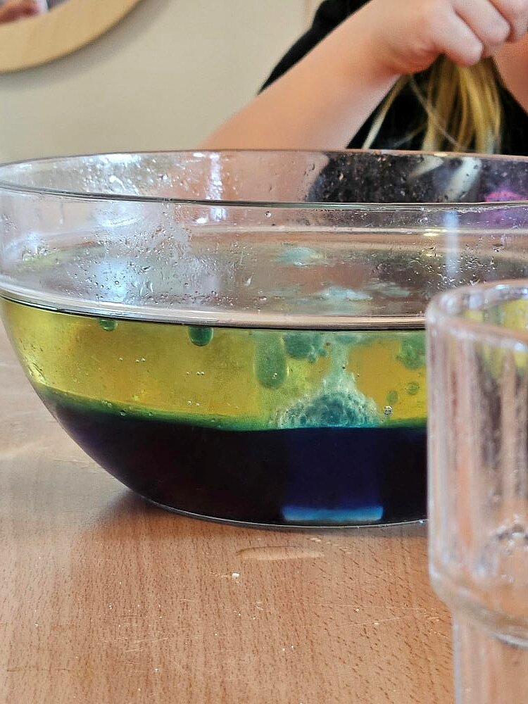 Ein Kind wirft eine Brausetablette in eine Glasschüssel, das bereits eine Schichtung aus farbigen Flüssigkeiten (grün, gelb und blau) enthält. Das Experiment findet auf einem Holztisch statt.